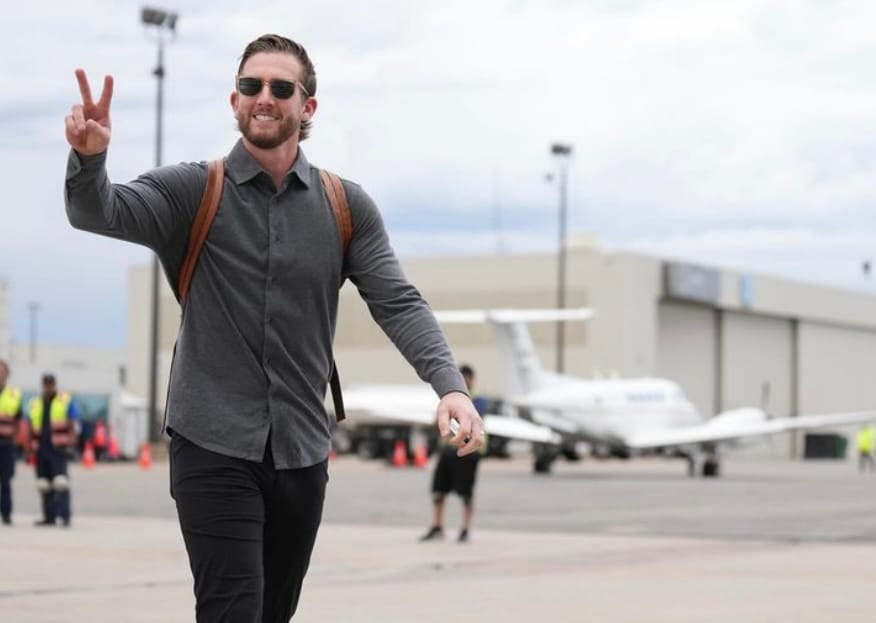 In the background are airplanes. McMahon wears a gray long-sleeved shirt and dark pants.